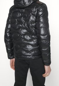 Doudoune noire avec une finition brillante, ornée de motifs en vagues matelassés et d'une capuche. Portée sur un pantalon noir, montrant la vue de dos.