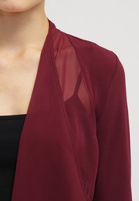 Burgundský oděv s průhledným, lehkým panelem. Má hladkou texturu a otevřený přední design, který vytváří efekt vrstvení.