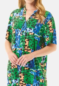 Camisa de manga corta en un vibrante diseño de patrones verdes, azules, naranjas y negros. Cuenta con cierre de botones en la parte delantera y un corte relajado, mostrando un diseño mixto.