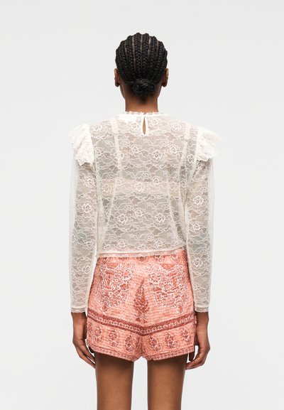 Femme aux cheveux tressés portant un chemisier blanc en dentelle à manches longues avec motifs floraux et un short taille haute rose à motifs, vue de dos.