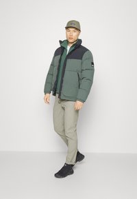 Puffjakke i mørkegrøn med sorte detaljer, med lynlås, høj krave og to sidelommer. Båret med beige bukser og sorte sko.