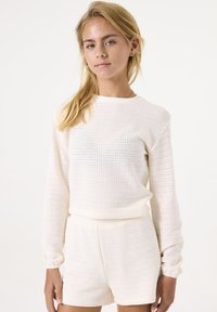 Weißer gestrickter Pullover mit langen Ärmeln und rundem Ausschnitt, kombiniert mit passenden hochgeschnittenen Shorts, die ein strukturiertes Offengewebe aufweisen.
