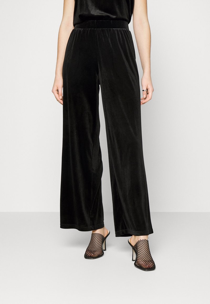 Femme portant un pantalon ample en velours noir et des mules en maille noire à talons hauts, debout devant un fond uni.