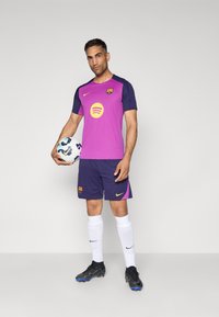 Violetinė ir tamsiai mėlyna futbolo apranga su logotipu, trumpomis rankovėmis ir atitinkančiomis šortais. Žaidėjas laiko raštuotą futbolą, vilkėdamas baltas kojines ir juodus sportbačius.