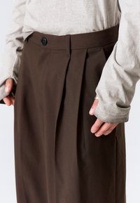 Pantalon plissé marron avec une ceinture plate et une fermeture par un seul bouton noir. Tissu texturé avec une coupe droite et décontractée.