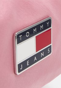 Różowa torba z prostokątnym logo Tommy Jeans, zawierającym biały kwadrat i czerwony prostokąt, podkreślona srebrnymi detalami i ciemnoniebieskim tłem.