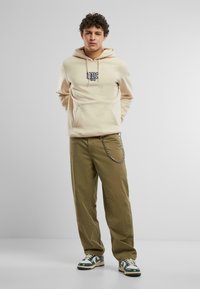 Felpa crema con tasca frontale e logo ricamato, abbinata a pantaloni verde oliva con un accento a catena, completata da sneakers multicolori.