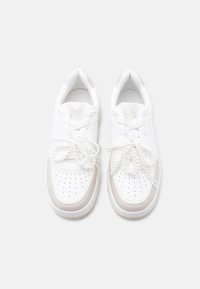 Baskets blanches avec panneaux texturés, embout perforé, accents crème et détails de lacets nacrés. Matériaux synthétiques et textiles lisses.