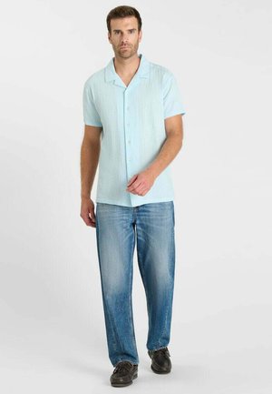 Mann trägt hellblaues kurzärmeliges Hemd mit Knöpfen, blaue Jeans und schwarze Schuhe, steht vor einem schlichten hellen Hintergrund.