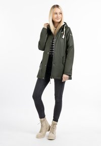 Olivgrön vattentät jacka med huva, tryckknappar och dragkedja, med en randig skjorta under, i kombination med svarta skinny jeans och beige stövlar.