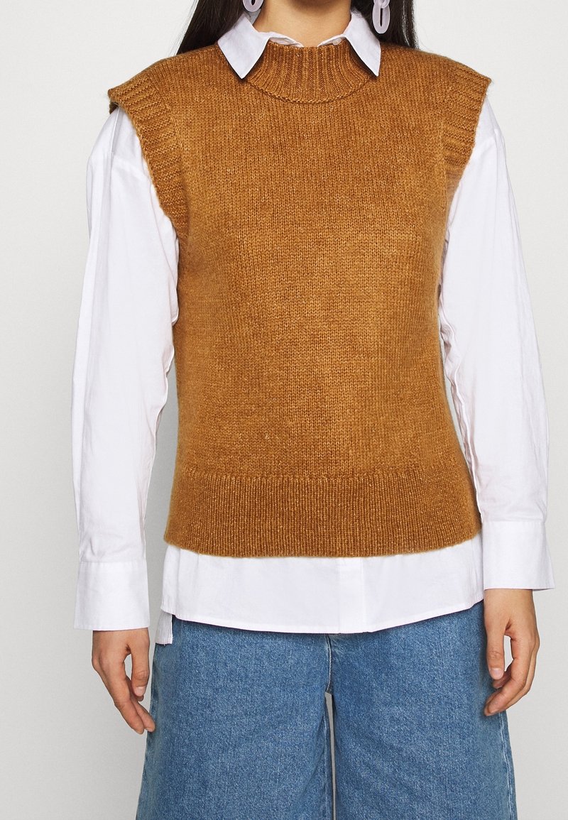 Pull sans manches en maille marron avec un col montant, doté d'un ourlet côtelé et d'une texture. Porté sur une chemise blanche à col. Coupe décontractée.