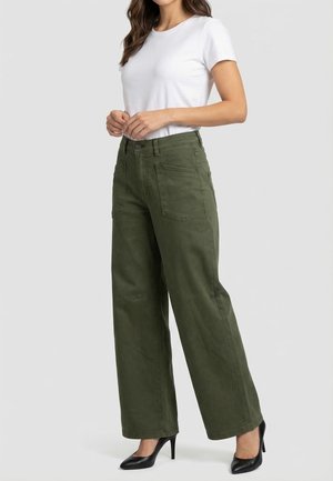 Vrouw die een wit T-shirt met korte mouwen, hooggetailleerde, wijd uitlopende olijfgroene broek en zwarte pumps draagt, staande tegen een effen achtergrond.