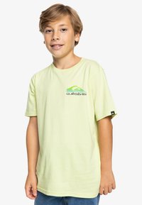 Camiseta de algodón amarillo claro con mangas cortas, que presenta un logo de Quiksilver en verde y negro en el lado izquierdo del pecho y una etiqueta negra en la manga.