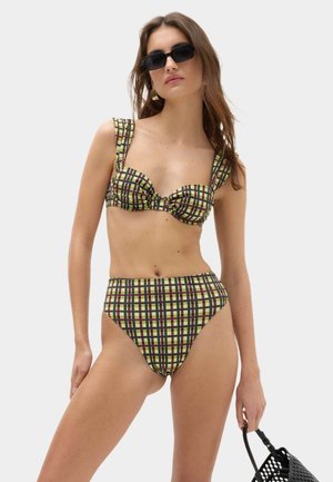 Kvinde iført grøn og brun ternet højhalset bikini med brede stropper og sorte rektangulære solbriller, der holder en sort perforeret håndtaske.