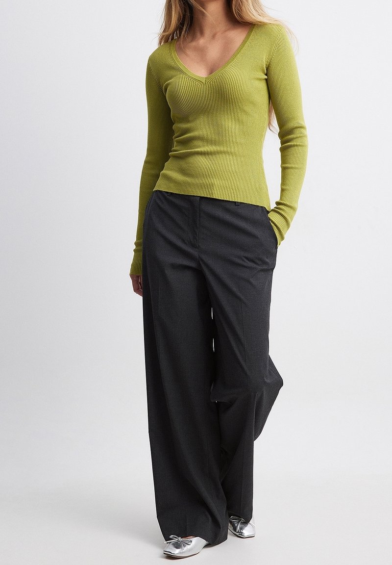 Donna con addosso un maglione a coste verde lime con scollatura a V, pantaloni a gamba larga grigio scuro e ballerine silver a punta, in piedi con una mano in tasca.