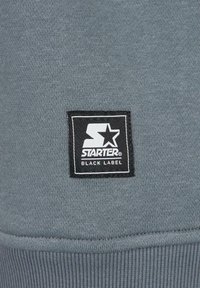 Patch du logo Starter Black Label sur tissu tricoté gris, affichant le nom de la marque avec un motif étoile en noir et blanc.
