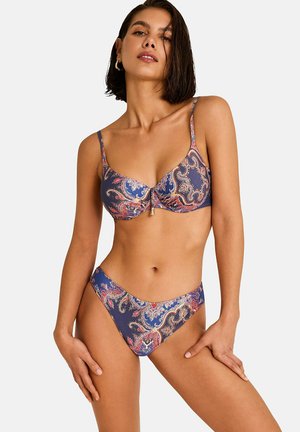 Femme portant un bikini deux pièces à motif cachemire dans des tons bleus, roses et beiges, posant avec une main sur la hanche.