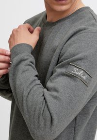 Sudadera gris con cuello redondo y puños acanalados. Presenta una etiqueta texturizada "Solid" cosida en la manga izquierda. Tejido suave con un corte relajado.