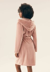 Różowy, pluszowy szlafrok z kapturem, z długimi rękawami, paskiem w talii oraz miękką fakturą. Widok z tyłu pokazuje długość do połowy łydki.