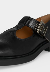 Pantof Mary Jane din piele lacuită neagră, cu o cataramă mare argintie și căptușeală interioară bej, văzut de aproape dintr-un unghi.