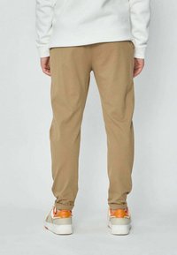 Personne portant un pantalon marron clair et des baskets blanches avec des accents orange, se tenant de dos contre un fond clair.