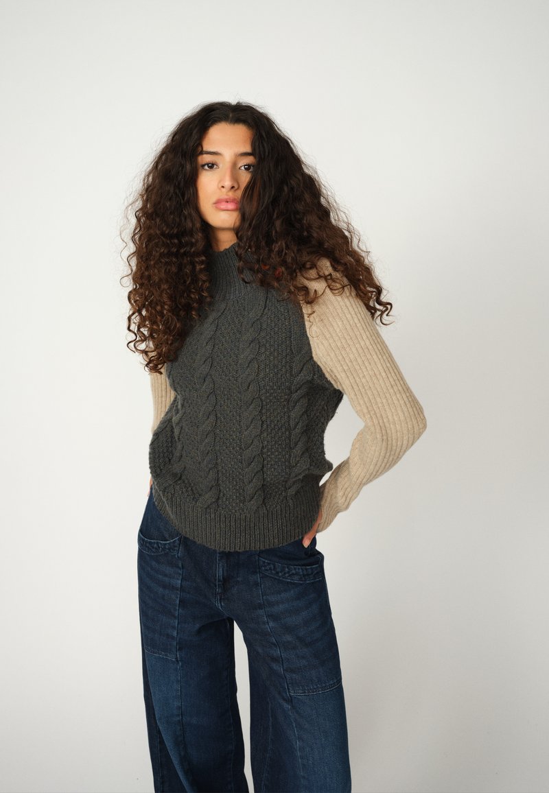 Der Pullover hat ein kabelgestricktes Design in Dunkelgrün mit einem gerippten beigen Ärmel. Kombiniert mit dunkelblauen weit geschnittenen Jeans. Strukturierte Oberfläche.