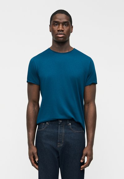 Man standing facing forward wearing a teal short-sleeve t-shirt and dark blue jeans against a plain light background.