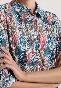 Camisa de botones con patrón floral sobre una base blanca. Presenta diseños en azul, rojo y marrón. La tela parece ligera y tiene mangas cortas.