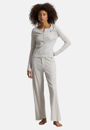 Femme aux cheveux bouclés portant un haut blanc à manches longues et un pantalon large à petits motifs floraux, debout pieds nus avec une main sur la hanche.