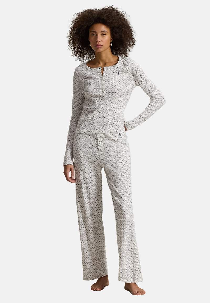 Vrouw met krullend haar, gekleed in een wit shirt met lange mouwen en wijde broek met kleine bloemenprint, staat blootsvoets met één hand op haar heup.