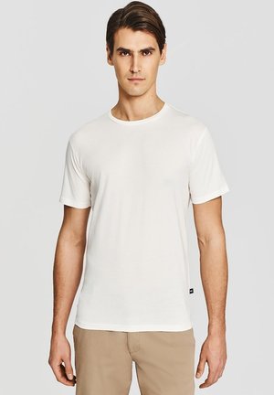 Młody mężczyzna z krótkimi brązowymi włosami, ubrany w gładką białą koszulkę z krótkim rękawem i beżowe spodnie, stojący na jasnoszarym tle.