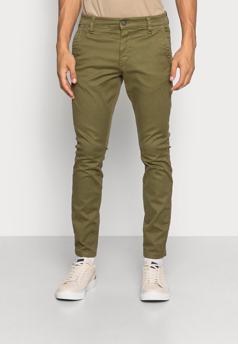 Homme portant un pantalon vert olive coupe slim et des baskets blanc cassé, debout sur un sol gris clair devant un mur blanc uni.