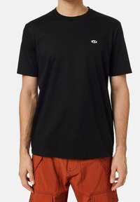 Černé tričko s krátkým rukávem a kulatým výstřihem, malé bílé logo na hrudi, v kombinaci s oranžovými cargo šortkami s viditelnými švy.