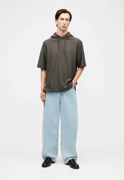 Νέος άνδρας με χαλαρό σκούρο πράσινο κοντομάνικο φούτερ με κουκούλα, φαρδιά ανοιχτό μπλε τζιν και μαύρα παπούτσια, στέκεται με το ένα χέρι στην τσέπη.