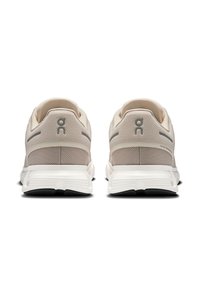 Beige athletic shoes with a mesh upper, reinforced heel, and white midsole. Gray accents and branding detail on the back. Black outsole.