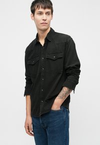 Czarna koszula dżinsowa z dwoma przednimi kieszeniami, zapinana na guziki, z podwiniętymi rękawami. Model ma na sobie niebieskie jeansy, na jego ramieniu widać tatuaż.