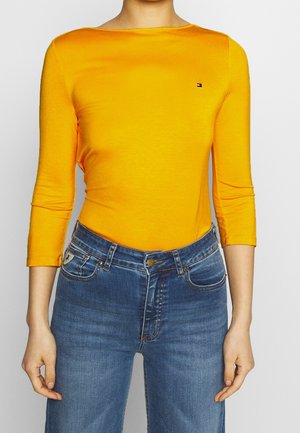 Kvinde iført en tætsiddende gul langærmet top med et lille logo på brystet og blå high-waisted jeans.