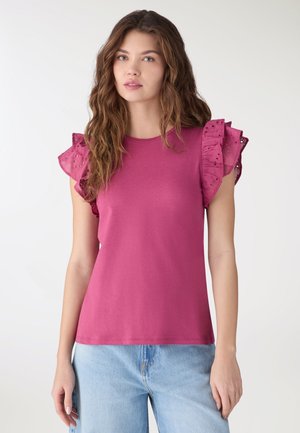 Donna con capelli lunghi e ondulati indossa una maglia rosa a maniche corte con dettagli a volant traforati e jeans azzurri, in piedi davanti a uno sfondo neutro.