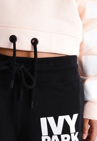 Felpa rosa chiaro tagliata con stringhe nere abbinata a pantaloni della tuta neri con logo bianco "IVY PARK" e vita a coste.