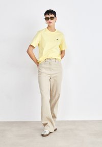 Camiseta de algodón amarilla de manga corta, combinada con pantalones de pierna ancha beige claro. Incluye zapatillas plateadas con suelas marrones y gafas de sol redondas.