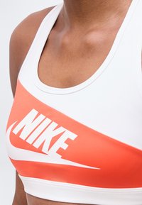 Κοντινό πλάνο ατόμου που φοράει λευκό και πορτοκαλί αθλητικό σουτιέν με έντονο λευκό λογότυπο Nike και τη χαρακτηριστική γραμμή swoosh στο στήθος.