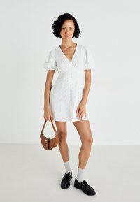 Biele krátke šaty s volánovými rukávmi a kvetinovým vzorom, s hlbokým V-krkom a zapínaním na gombíky vpredu. Nosené s čiernymi topánkami a hnedou kabelkou.