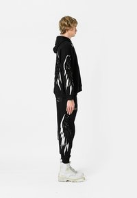 Sudadera negra y pantalones con diseños de alas blancas y con plumas. El modelo lleva botas blancas. La tela tiene una textura suave y un ajuste relajado.