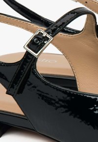 Zwarte patenten leren sandaal met kruisbandjes en zilveren vierkante gesp. Bevat een beige inlegzool en een lage hak voor stabiliteit.