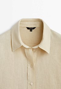Chemise en lin beige à boutons, col classique et étiquette de marque visible à l'intérieur de l'encolure.