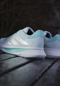 Helle blaue Sportschuhe mit einem strukturierten Obermaterial und drei silbernen Streifen; verfügen über eine glatte weiße Sohle und Akzentdetails in dunklerem Blau.