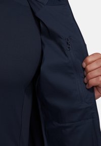 Navy jacket interior features a textured fabric with a zippered pocket. A person’s hand is positioned near the pocket, grasping the fabric.