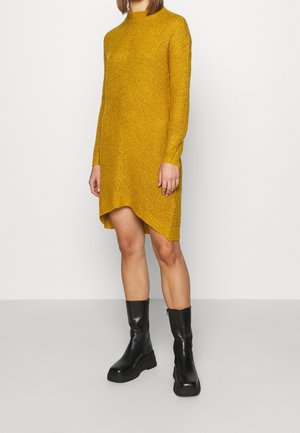 Senfgelbes Strickkleid mit langen Ärmeln und asymmetrischem Saum. Getragen mit schwarzen Stiefeletten mit klobiger Sohle.