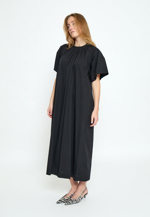 Frau steht in einem locker sitzenden schwarzen Midikleid mit kurzen Ärmeln und trägt schwarz-weiß gemusterte spitze Slingback-Pumps mit Zebradruck.