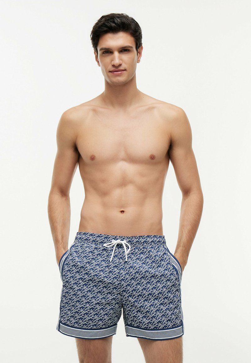 Jeune homme aux cheveux foncés, torse nu, portant un short de bain bleu à motifs avec un cordon blanc, les mains dans les poches.